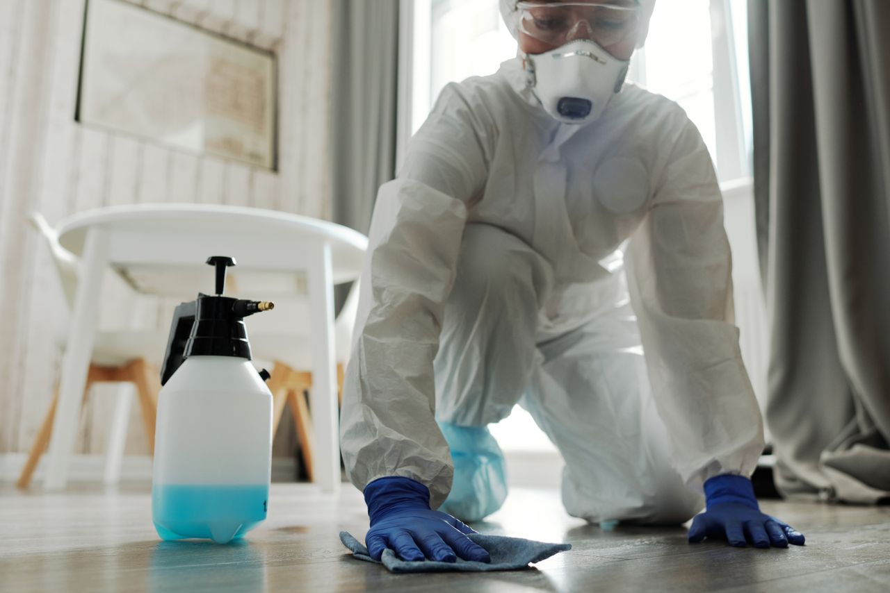 A person wearing protective equipment cleans an indoor floor using disinfectant spray and a cloth.